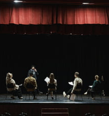 group of people in chairs on a stage reading texts in their laps